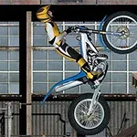 Motorkář v žlutém oblečení dělá trik na motorce před industriálním pozadím s okny a bednami